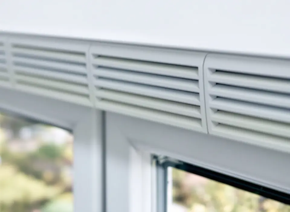 Een wit ventilatierooster boven in een wit kunststof kozijn, van binnen gezien